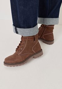 Bottes chelsea en cuir marron avec lacets et fermeture éclair latérale, portées avec un jean en denim foncé retroussé sur un sol clair.