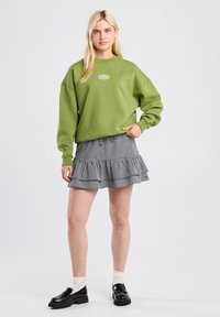 Felpa verde oversize con collo a girocot  a costine, abbinata a una gonna con volante in gessato bianco e nero e mocassini in pelle lucida.