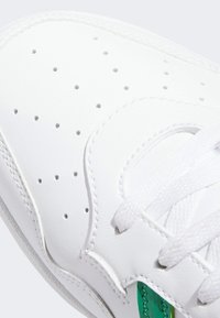 Zapatilla blanca de cuero con puntera perforada, cordones blancos planos y un detalle verde cerca de los ojales. Textura lisa con detalles de costura.