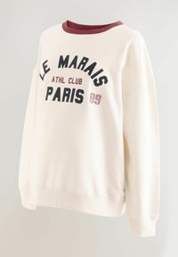 Sudadera de color crema con cuello redondo burdeos, que presenta un texto negro en negrita "LE MARAIS ATHL CLUB PARIS 89" en la parte frontal. Textura suave.