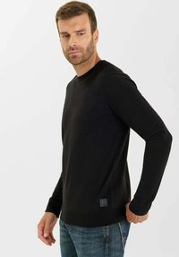 Uomo con capelli corti e castani e barba, indossa un maglione nero a maniche lunghe e jeans blu, in piedi davanti a uno sfondo chiaro e uniforme.