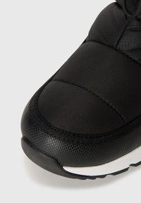 Sapatilha desportiva preta com um cabedal têxtil suave e mate, detalhes acolchoados e uma sola de borracha texturizada com uma faixa branca.