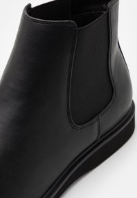 Botín Chelsea de cuero negro con panel lateral elástico y suela de goma baja sobre un fondo blanco.