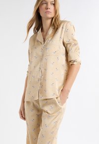 Kremowy zestaw piżamowy w kwiatowy wzór. Składa się z zapinanego na guziki topu z kieszonką oraz pasujących spodni, oba wykonane z gładkiej tkaniny.