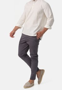 Mężczyzna w białej koszuli z długim rękawem, ciemnoszarych spodniach i beżowych espadrylach na płaskiej podeszwie, pozujący z jedną ręką w kieszeni i jedną nogą skrzyżowaną.