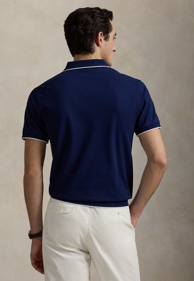 Μπλε ναυτικό πόλο με λευκή επένδυση στο γιακά και τα μανίκια, κατασκευασμένο από λείο ύφασμα· φορεμένο με ανοιχτόχρωμο παντελόνι, από πίσω.