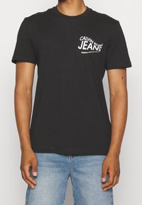 Black cotton t-shirt with short sleeves, featuring a white "Calvin Klein Jeans" logo on the left chest and a classic round neckline.
