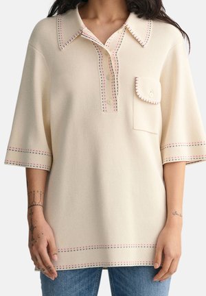 Femme portant un haut en tricot crème à manches courtes avec col, trois boutons, une poche poitrine et des détails de bordure cousus rouges et noirs.