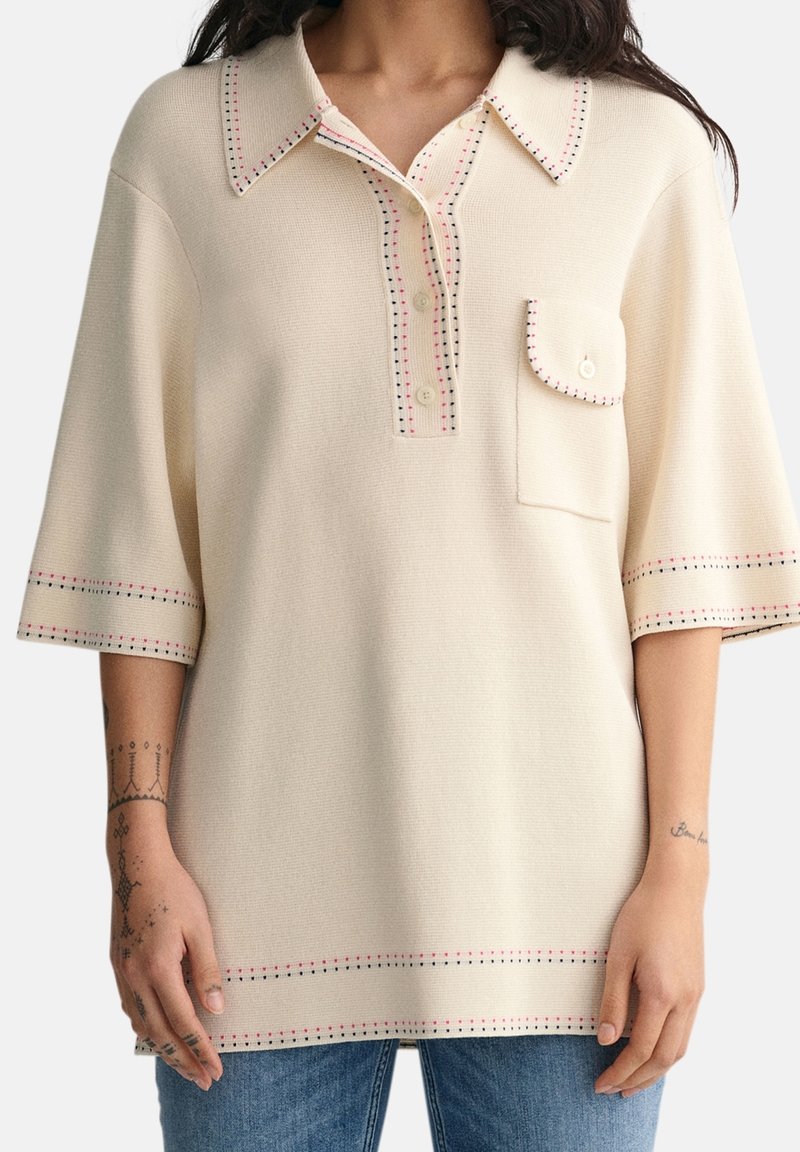 Femme portant un haut en tricot crème à manches courtes avec col, trois boutons, une poche poitrine et des détails de bordure cousus rouges et noirs.