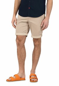 Beige fabric shorts with rolled cuffs, worn with bright orange slide sandals. Model stands with one hand on hip, showcasing casual outfit.