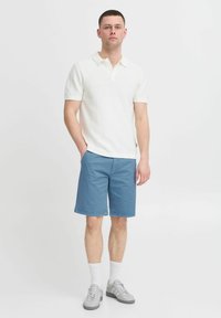 Polo shirt blanc texturé, shorts bleus, baskets grises et chaussettes blanches. La chemise possède un col classique et des manches courtes.
