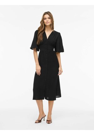 Žena stojaca, oblečená v čiernych midi šatách s gombíkmi vpredu a volánikovými rukávmi, v kombinácii s tmavými otvorenými sandálmi na podpätku, na bielom pozadí.