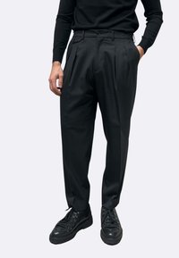 Pantaloni neri plissettati in tessuto liscio, con due tasche frontali e vestibilità sartoriale, abbinati a sneakers nere.