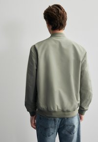 Licht olijfgroene bomberjack met een gladde textuur, geribbelde manchetten en zoom, en een ontspannen pasvorm, gecombineerd met lichtblauwe jeans.