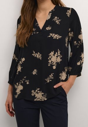 Woman wearing a black V-neck blouse with beige floral prints and three-quarter sleeves, paired with dark pants, hand in pocket.
