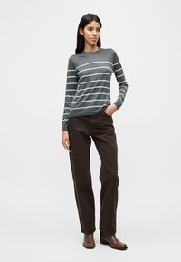 Ριγέ πουλόβερ μακρυμάνικο σε σκούρο γκρι με λευκές ρίγες· συνδυασμένο με καφέ παντελόνι και καφέ μπότες με τακούνι. Απλός λαιμός, στενή εφαρμογή.