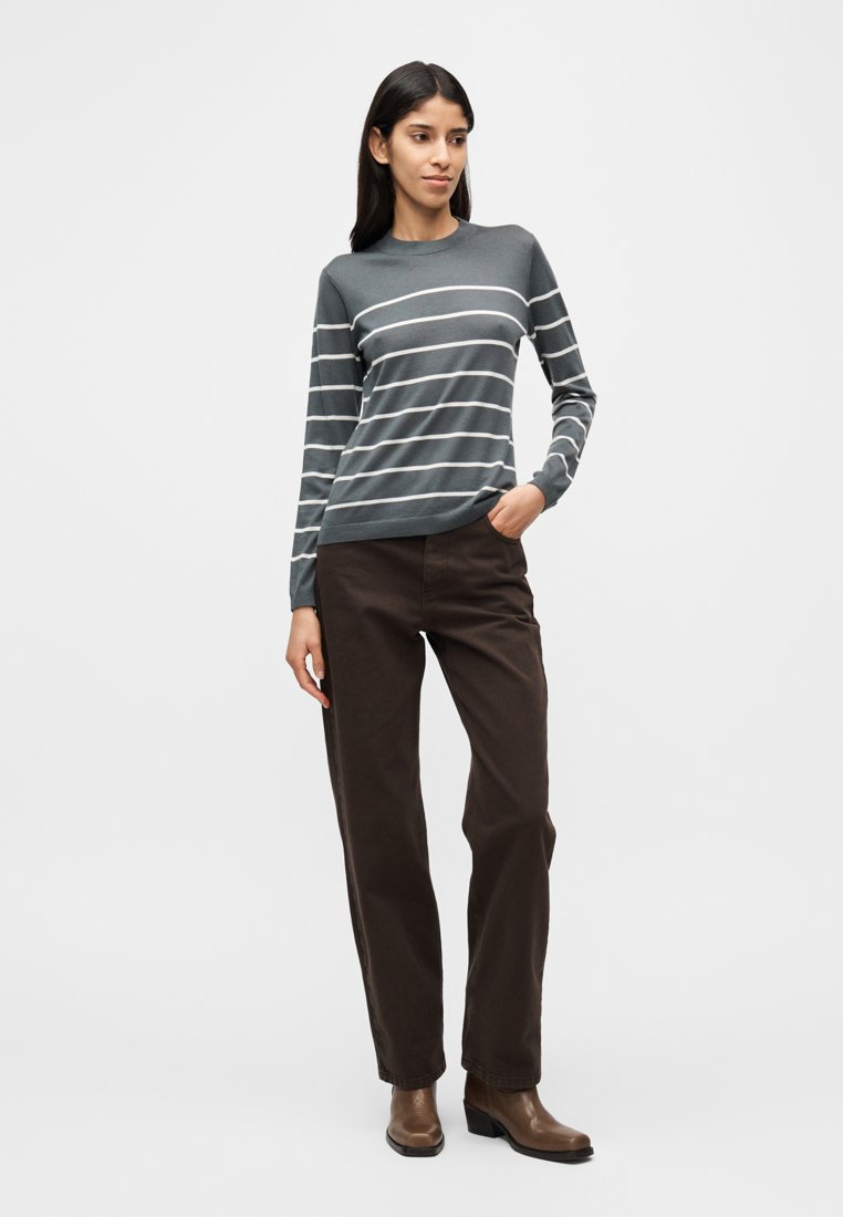 Pull à manches longues rayé gris foncé avec des rayures blanches ; associé à un pantalon marron et des bottes à talons marron. Encolure simple, coupe ajustée.