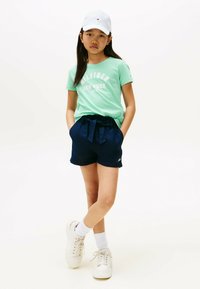 Camiseta de algodón verde claro con estampado "Tommy Hilfiger New York", combinada con pantalones cortos azul marino con cinturilla ajustable. Zapatillas blancas y gorra.