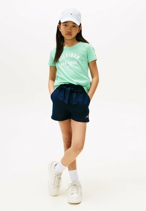 Jasnozielona bawełniana koszulka z nadrukiem "Tommy Hilfiger New York", w połączeniu z granatowymi szortami z wiązaniem w pasie. Białe sneakersy i czapka.