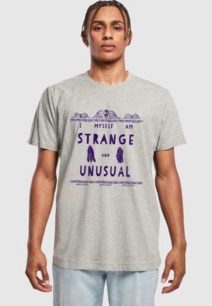 Grå bomulds t-shirt med lilla tekst, der siger "Jeg er selv MÆRKELIG og UDSÆT", med designelementer og accenter.