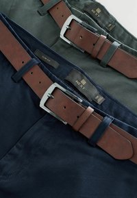 Uske slim-fit hlače u tamnozelenoj i mornarsko plavoj boji s smešnim kožnim remenima. Imaju srebrne metalne kopče i vidljive šavove. Unutra su pričvršćene etikete.