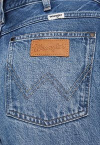 Tylnia kieszeń niebieskich jeansów z naszywką ze skóry i materiałową metką, obie z nazwą marki „Wrangler”.