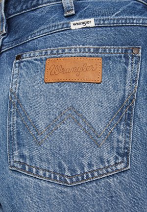 Zadný vrecko na modrých rifliach s koženou záplatou a textilnou nálepkou, obe zobrazujú značku „Wrangler“.