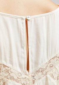 Krämfärgad blus med spetsdetaljer nertill och en liten knappstängning på baksidan, med en slitsöppning under.