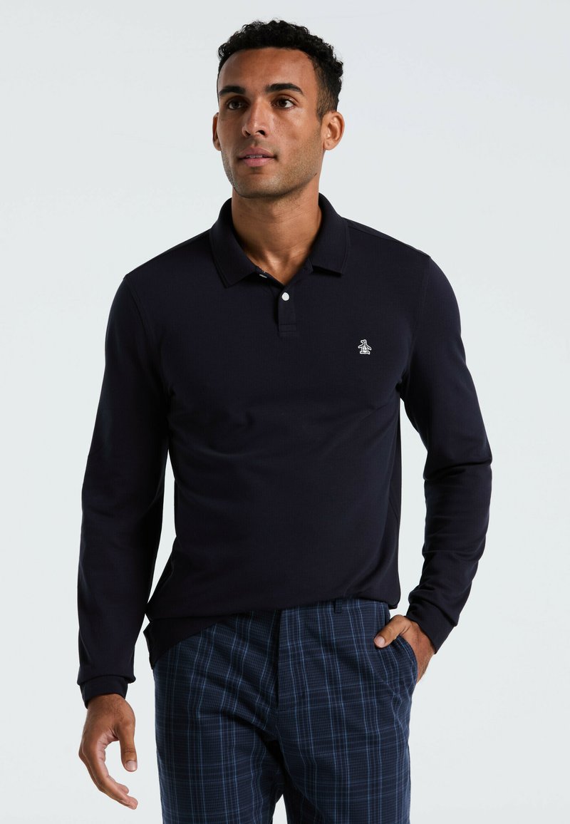 Marineblauw poloshirt met lange mouwen en een drie-knopen-knoopsluiting, voorzien van een klein logo op de borst. G gedragen met geruite broek in marine en blauw.