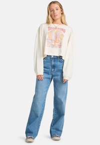 White cropped long-sleeve shirt with a colorful graphic design, paired with high-waisted light blue wide-leg jeans and gray sneakers.
