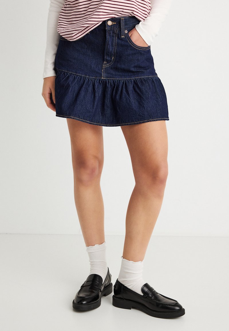 Άτομο που φοράει σκούρο μπλε τζιν μίνι φούστα με βολάν, λευκές κάλτσες, μαύρες μοκασίνες και ριγέ τοπ, με το ένα χέρι στην τσέπη.