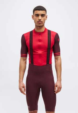 Άνδρας που φοράει κόκκινη και σκούρα μπορντό φανέλα ποδηλασίας με μαύρες τιράντες και ταιριαστά σορτς ποδηλασίας, στέκεται μπροστά σε απλό φόντο.