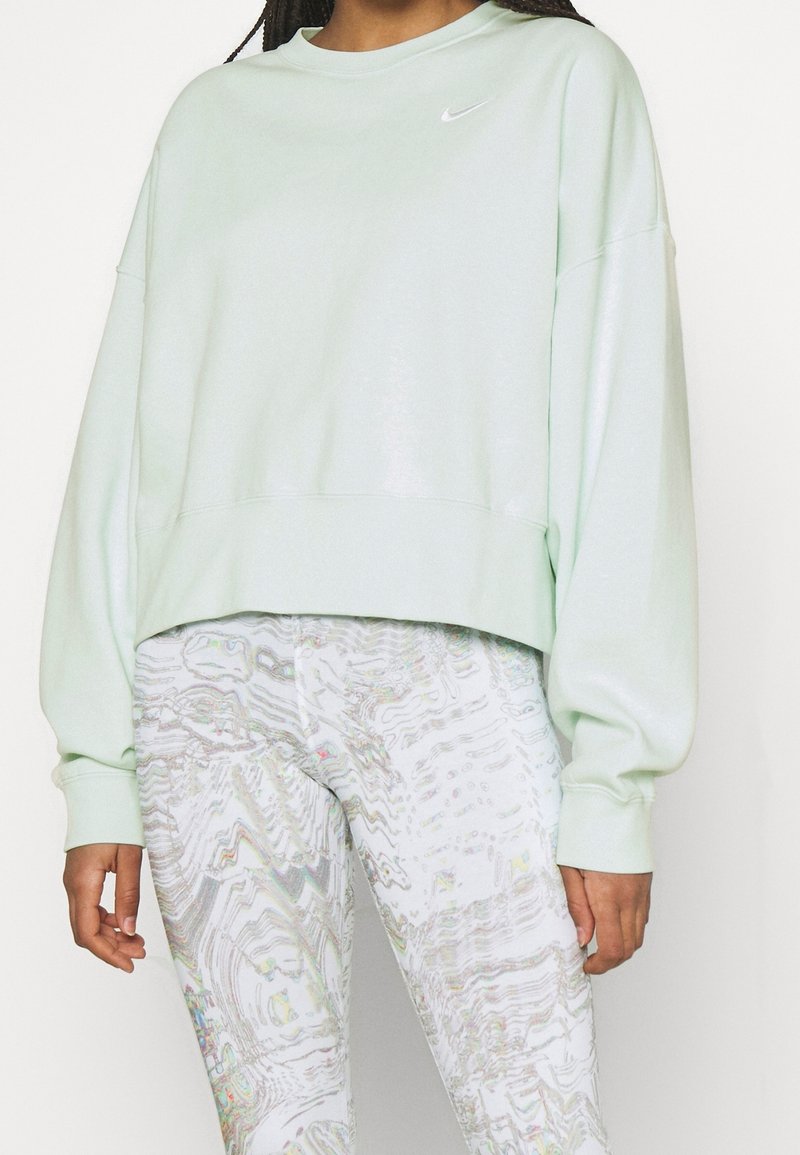 Femme portant un sweat-shirt Nike vert clair oversize et un legging blanc à motifs, debout devant un fond uni.