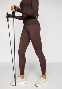 Braunes Langarmoberteil und passende Leggings aus dehnbarem Material. Schwarze Sportstöcke und hellfarbene Sportschuhe sind sichtbar.