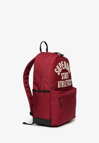 Bordeauxfarvet rygsæk med sort bund, frontlommelomme med lynlås, polstrede stropper og teksten "SUPERDRY STATE ATHLETICS" på hovedrummet.