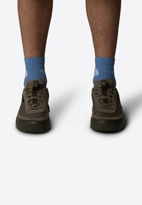 Zapatillas marrones con una parte superior de lona texturizada y suelas de goma negras, combinadas con calcetines de rayas azul claro que tienen un logo.