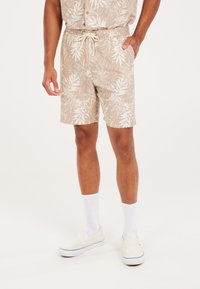 Pantalones cortos beige con un patrón de hojas blancas, cinturilla elástica con cordón, bolsillos laterales y un ajuste relajado. Combinados con calcetines y zapatos blancos.