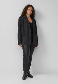 Mustriga musta bleiser koos lõigatud krae, sobib kokku sobivate mustade pükste ja pahkluu saapatega, luues stiilse ja struktureeritud silueti.