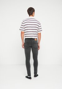 Jeans skinny grises con bolsillos traseros y una etiqueta marrón. Combinados con una camisa de manga corta blanca a rayas y zapatillas negras.