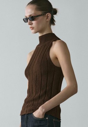 Donna che indossa occhiali da sole rettangolari scuri e una maglia smanicata marrone a coste con collo alto, mano in tasca, su sfondo neutro.