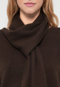 Mulher usando um cachecol de malha castanho-escuro enrolado em volta do pescoço e um suéter a condizer, com lábios e queixo visíveis.