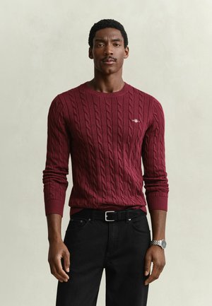 Mann trägt einen burgunderfarbenen Kabelstrickpullover, schwarze Hose mit Gürtel und eine silberne Armbanduhr, steht vor einem schlichten hellen Hintergrund.