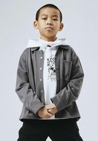 Chaqueta gris de botones sobre una sudadera blanca con gráfico; cuenta con un bolsillo frontal y un corte relajado. Pantalones negros completan el atuendo. Fondo liso.