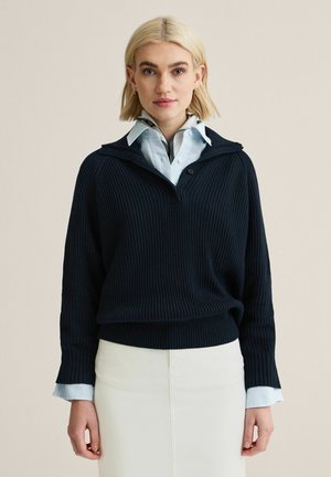 Vaaleahiuksinen nainen, jolla on päällään laivastonsininen ribbineulospusero vaaleansinisen kauluspaidan ja valkoisen hameen päällä, beigen taustan edessä.