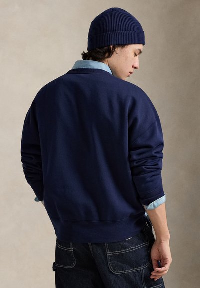 Granatowa bluza dresowa o luźnym kroju, z prążkowanymi mankietami i okrągłym dekoltem, noszona na jasnoniebieskiej koszuli z kołnierzykiem, do tego dopasowana czapka beanie.