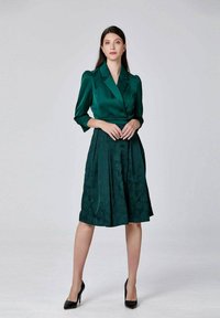 Femme se tenant debout vêtue d'un haut en satin vert foncé de style blazer et d'une jupe texturée jusqu'aux genoux assortie, avec des talons noirs, sur un fond uni.