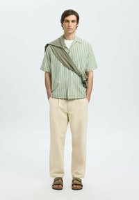 Camisa de manga corta a rayas verde claro, pantalones crema, sandalias caqui, con un suéter gris drapeado sobre un hombro, de pie contra un fondo sencillo.