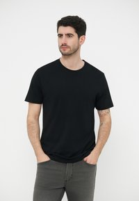 Νεαρός με κοντά σκούρα μαλλιά, φορώντας μαύρο κοντομάνικο μπλουζάκι και γκρι τζιν, με τα χέρια στις τσέπες, κοιτάζοντας ελαφρώς προς τα πλάγια.