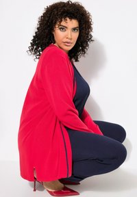 Roter Cardigan mit langen Ärmeln, offener Front, schrägem Saum und seitlichem Schlitz, getragen über einem navyfarbenen Oberteil und Hosen. Glänzende rote High Heels.