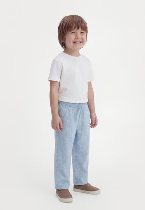 Uśmiechnięty młody chłopiec stojący, ubrany w białą koszulkę z krótkim rękawem, jasnoniebieskie spodnie i brązowe buty wsuwane na białym tle.